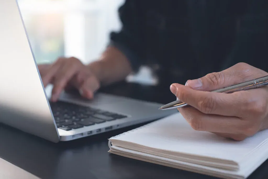 Mains sur un ordinateur portable, stylo au-dessus d’un carnet